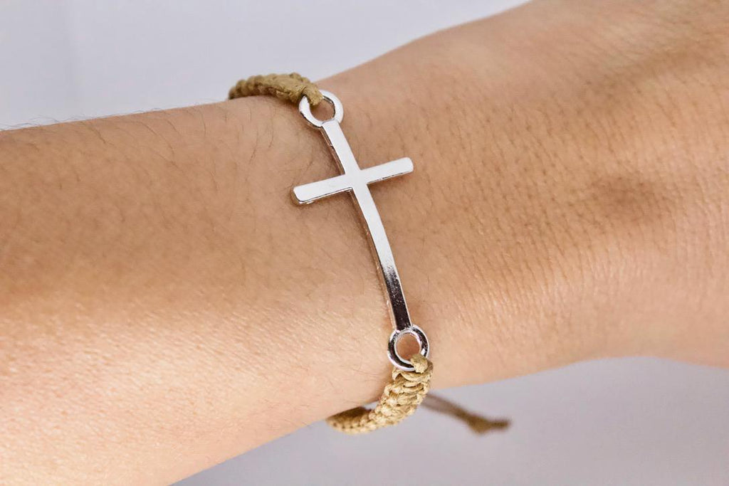 Silver cross bracelet on a wrist with a gold braided band against a neutral background