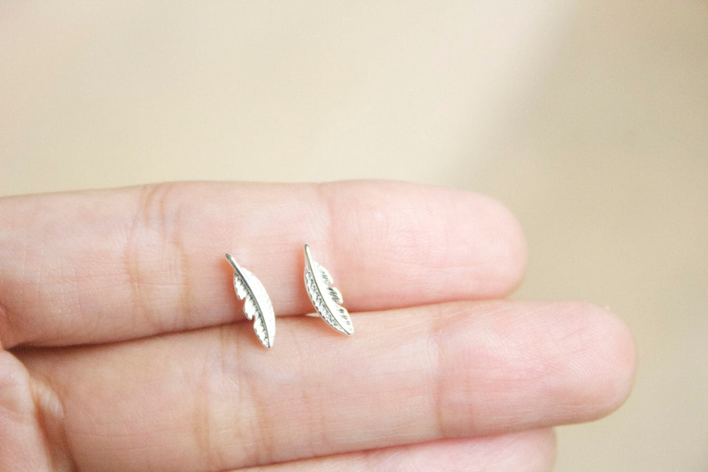 Silver feather-shaped earrings held between fingers against a blurred background