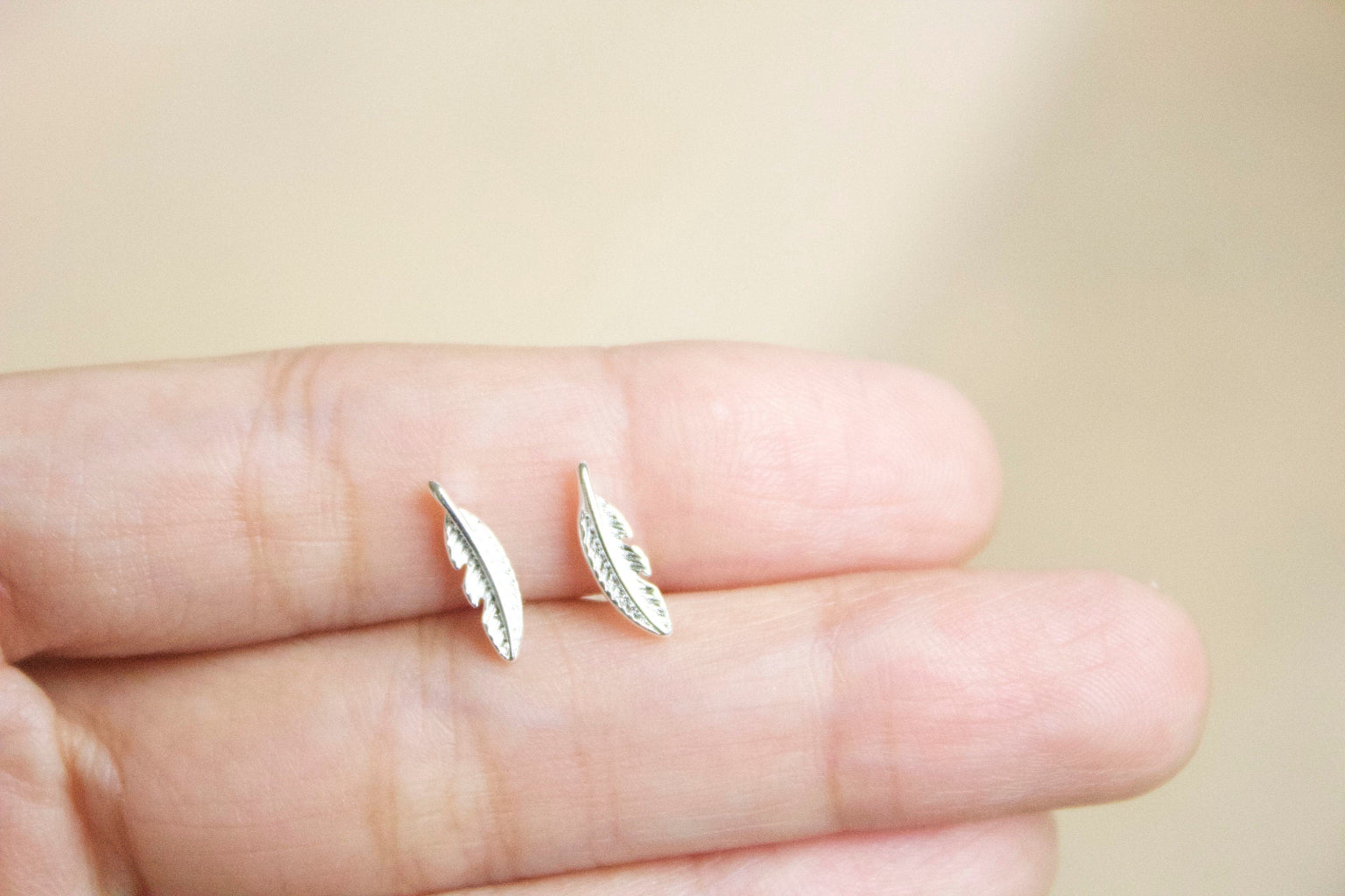 Silver feather-shaped earrings held between fingers against a blurred background