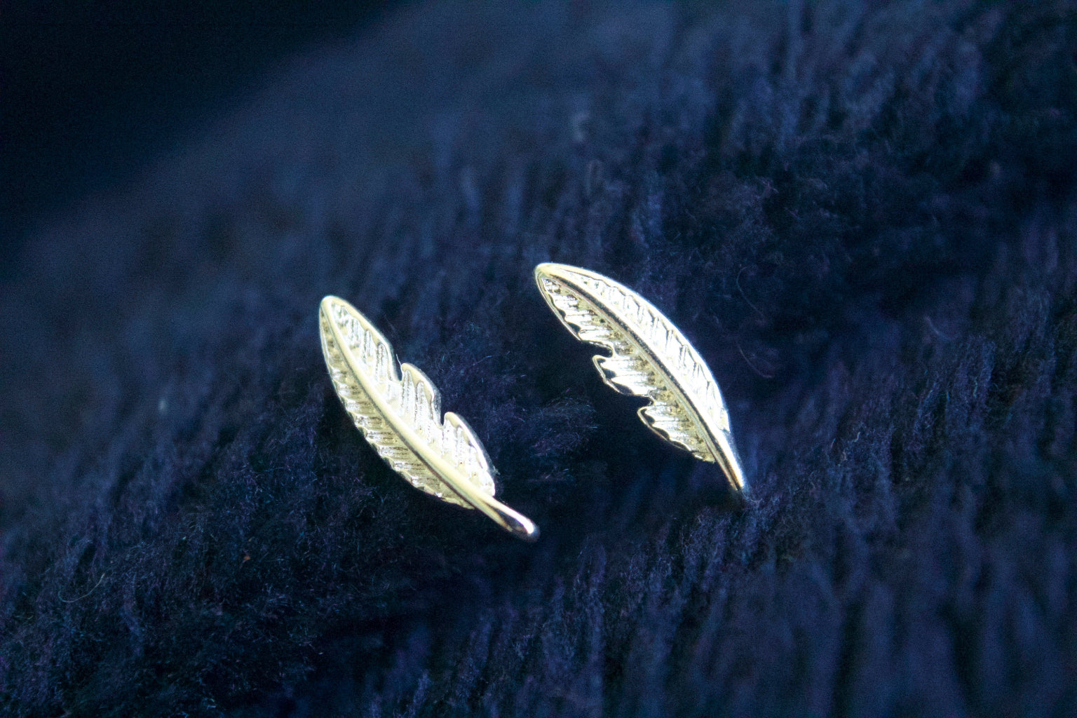 Silver feather earrings on a dark blue textured fabric background