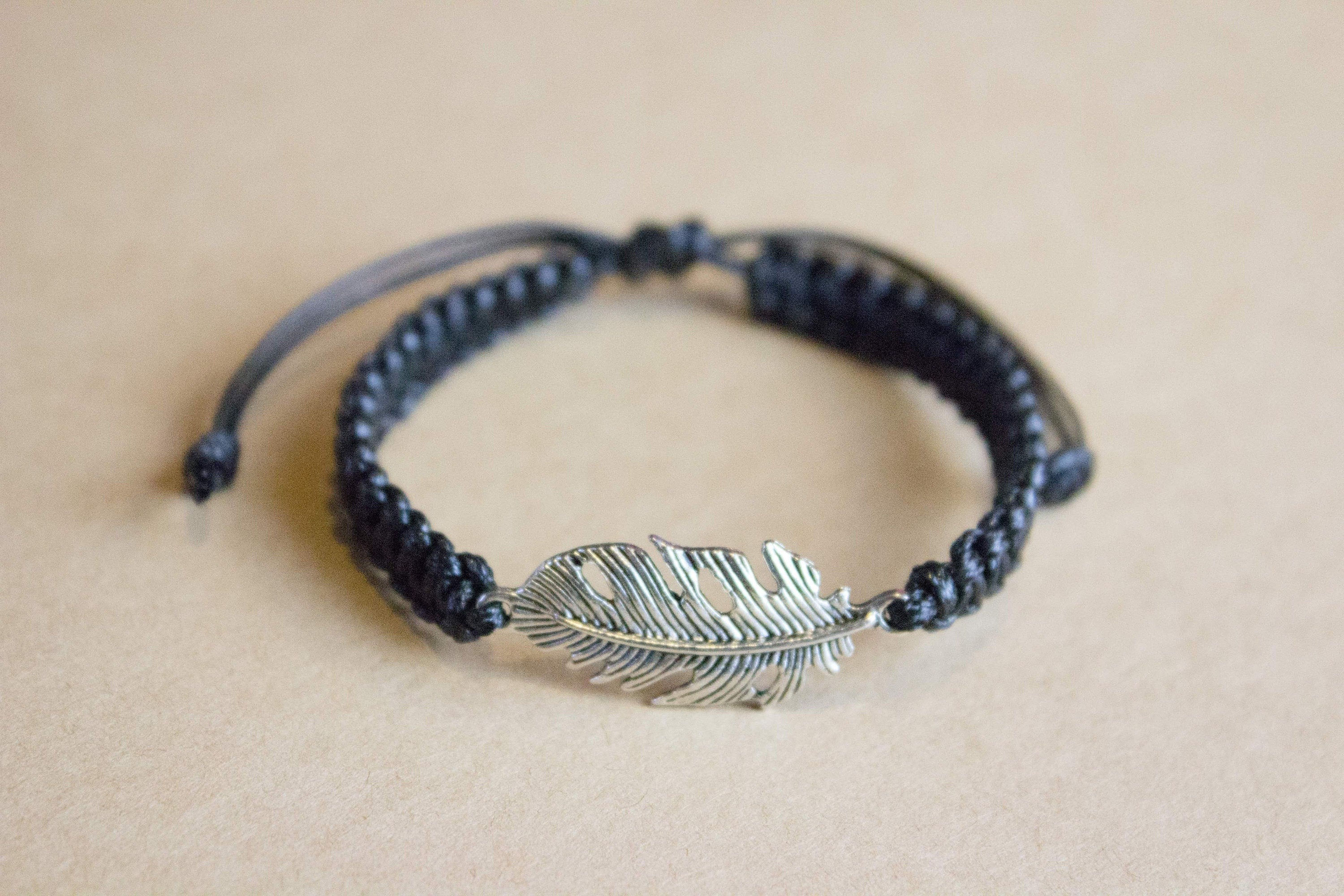 Black braided bracelet with a silver feather charm on a beige background