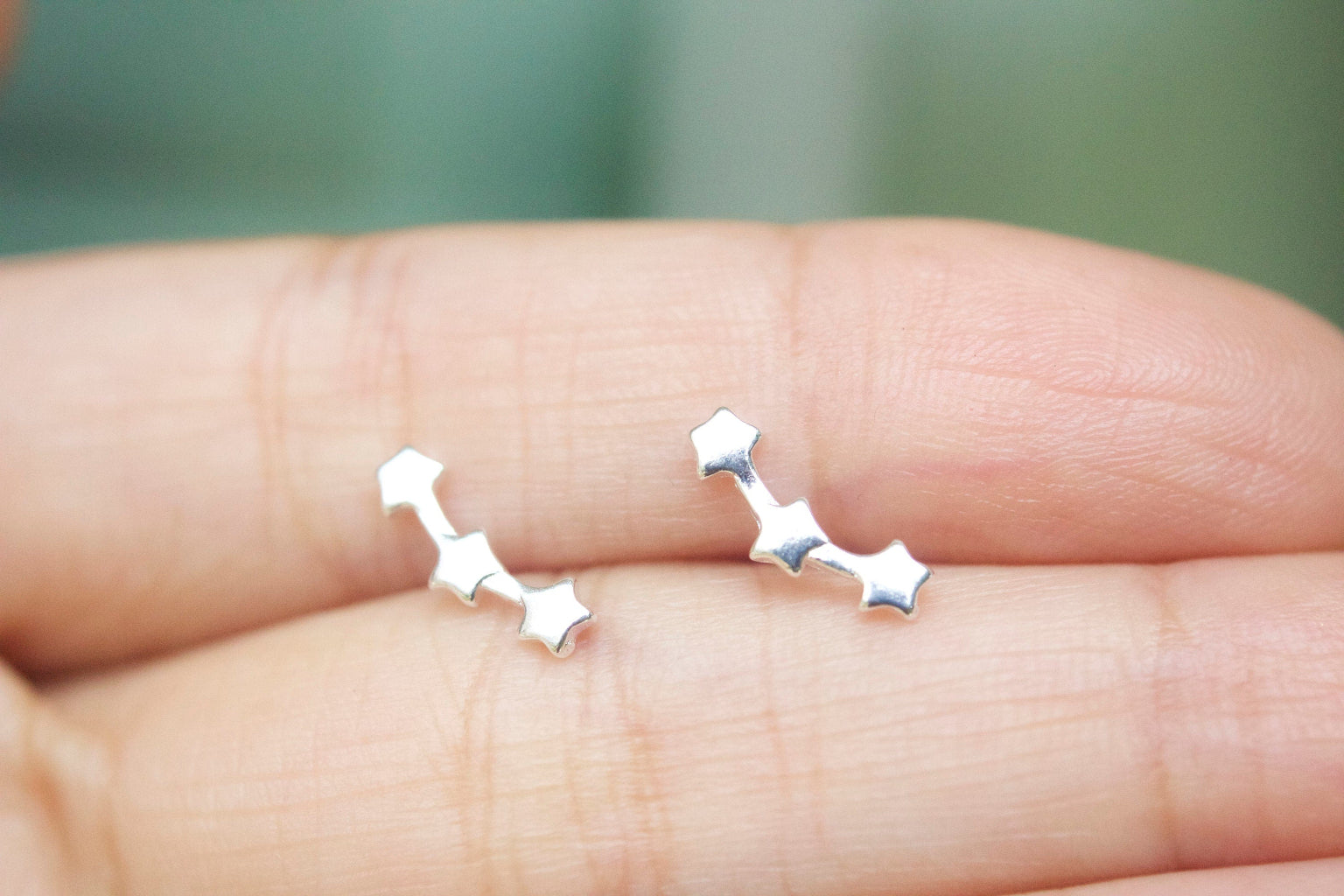 Silver star-shaped earrings held between two fingers against a blurred green background