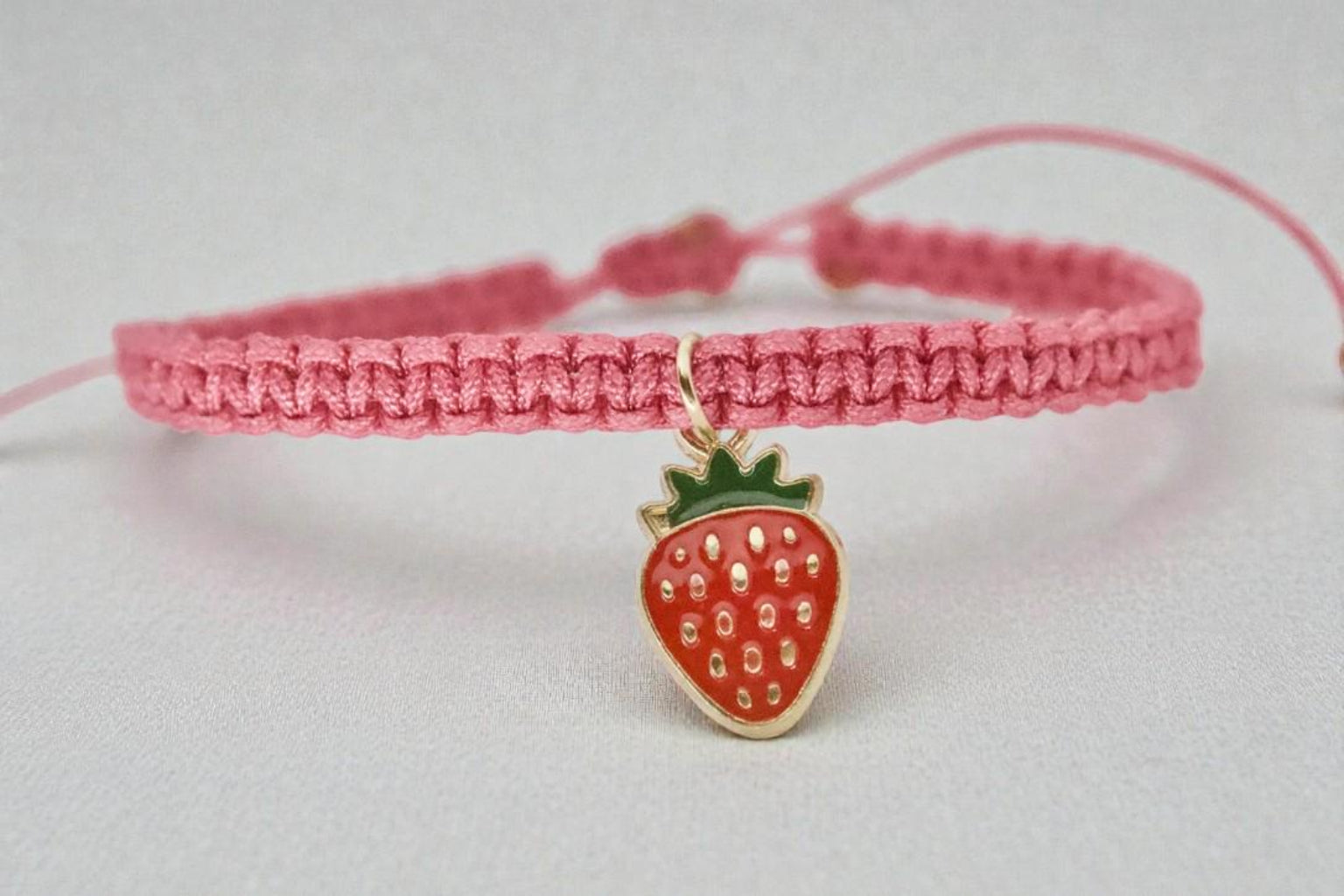 Pink braided bracelet with a strawberry charm on a light gray background
